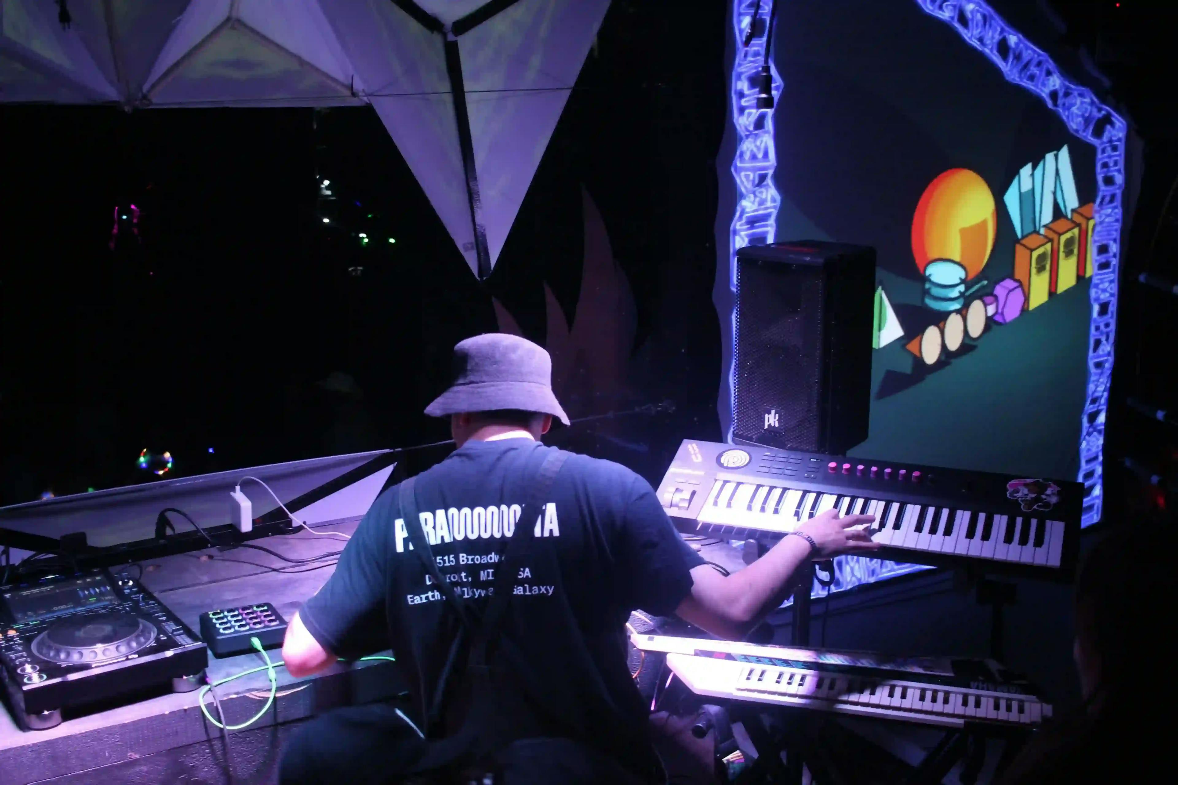 Rear view of a DJ performing to a crowd at Asgard stage, under a tent with lighting equipment.