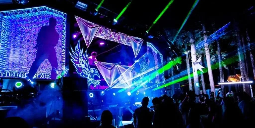 A laser-lit DJ stage at night in a forest with a crowd.
