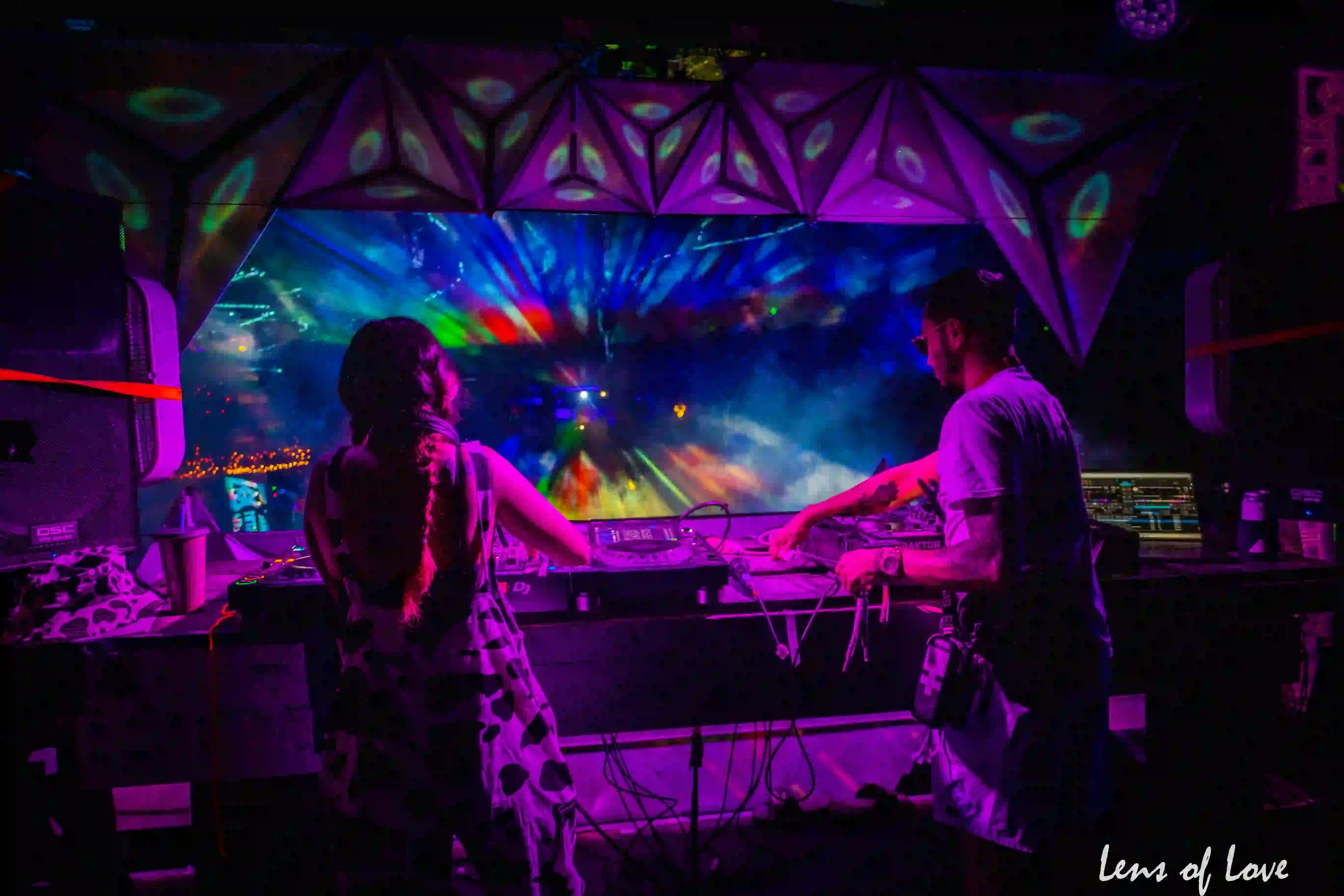 DJs at a neon-lit console with a vibrant visual display at a nighttime event.
