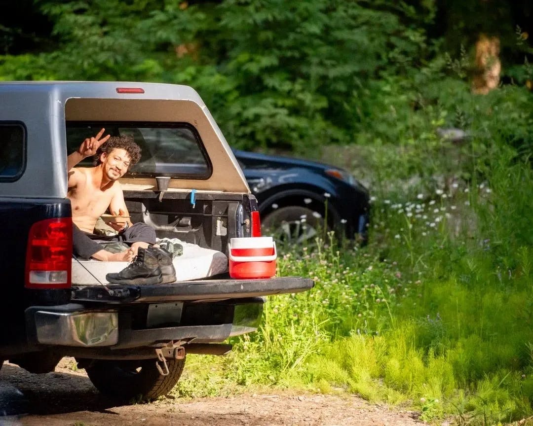 Man giving a peace sign while sitting in the bed of a pickup truck with camping gear.