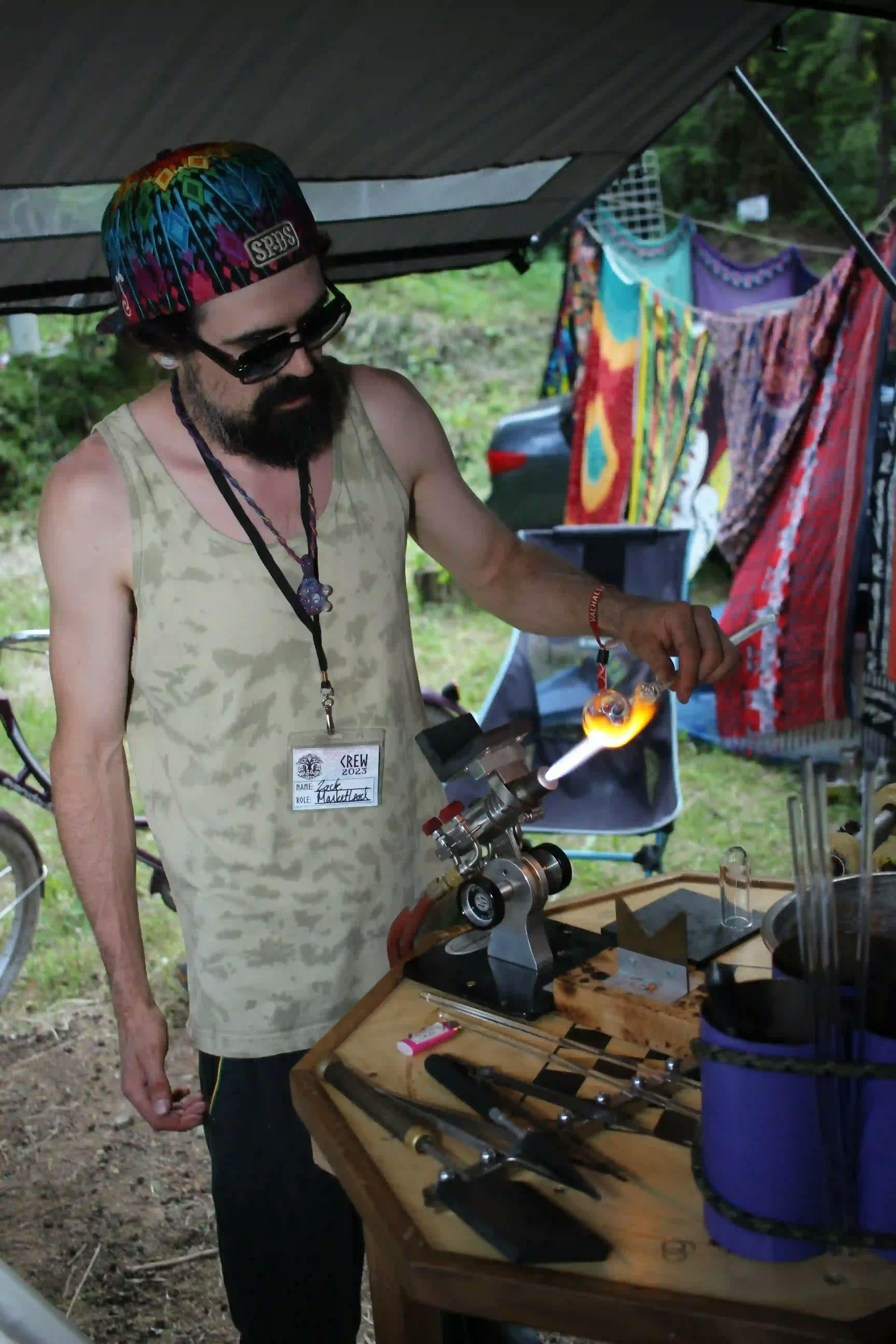 A man operating a glassblowing torch at a craft station with tools, wearing a colorful hat and a crew badge.