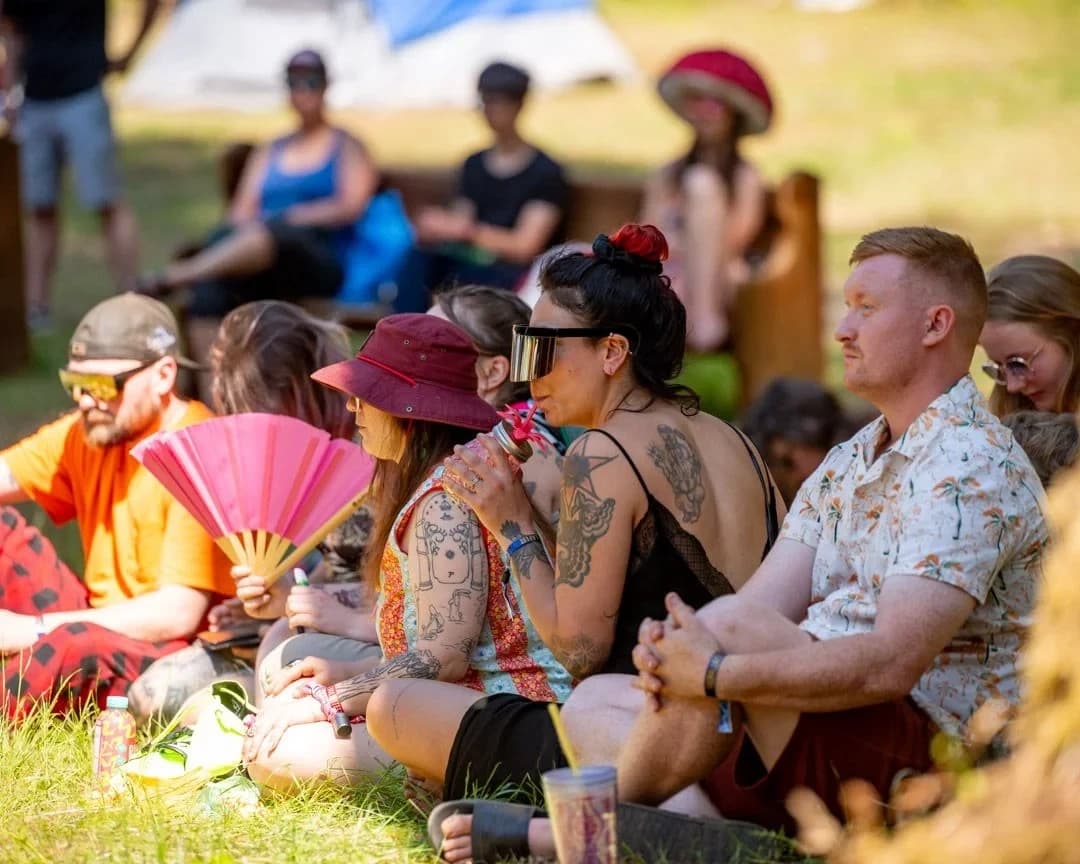 Festival attendees sitting on the grass, relaxing and listening to a speaker, with a focus on casual summer attire and accessories.