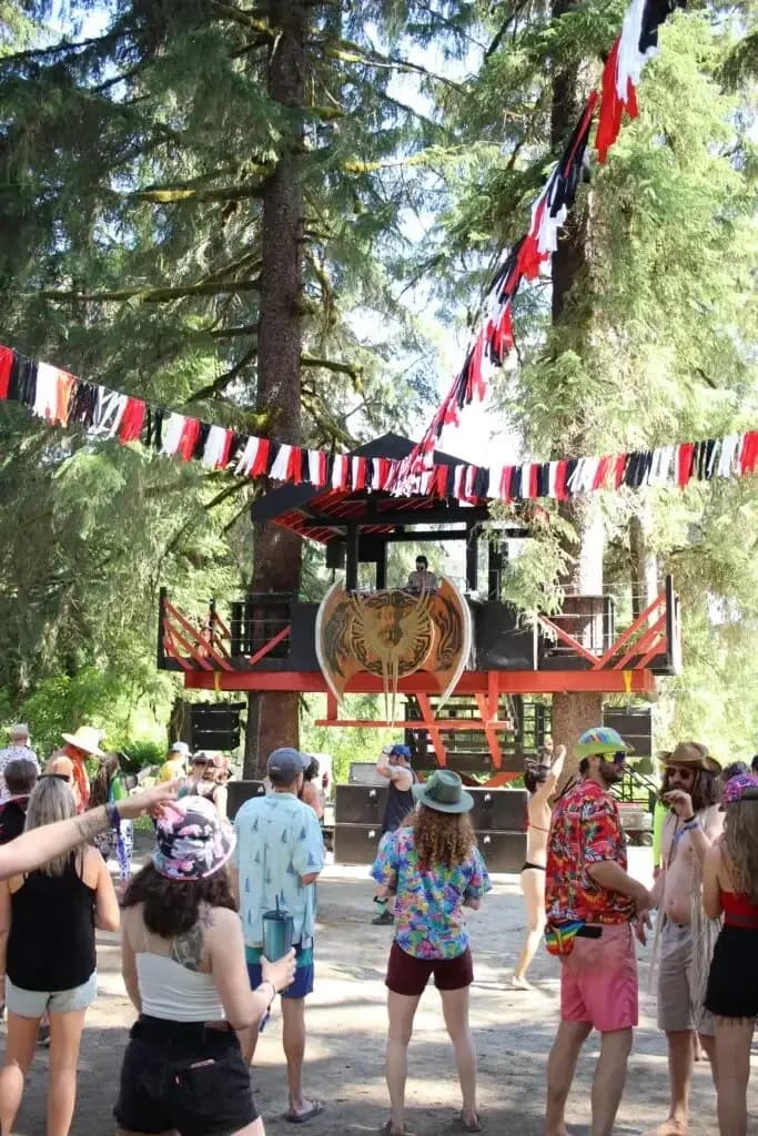 Festival goers dancing under bunting flags in a forest clearing.