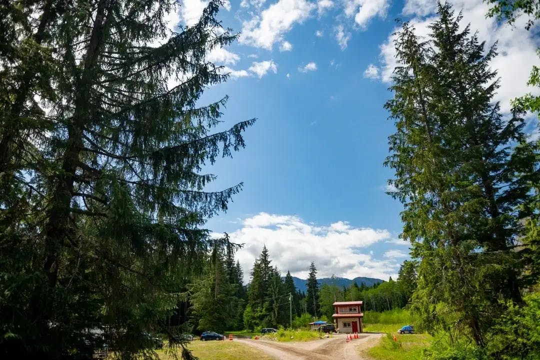 Entrance to ValhallaFest with a Gate House along a gravel path, surrounded by tall evergreens under a sunny, cloud-speckled blue sky.
