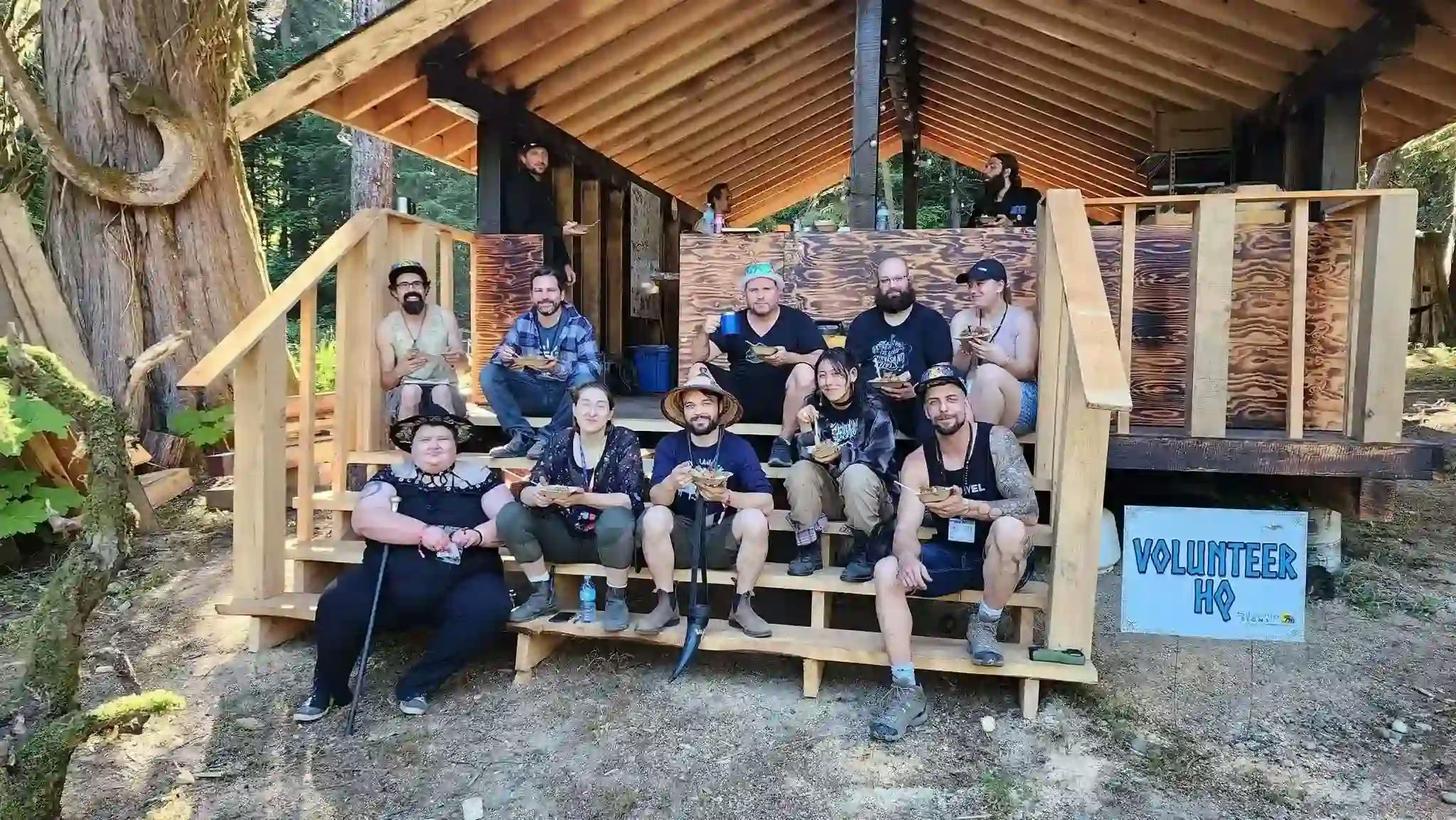 A group of smiling volunteers from Valhallafest 2023 gathered on steps of a wooden cabin in a forest setting.
